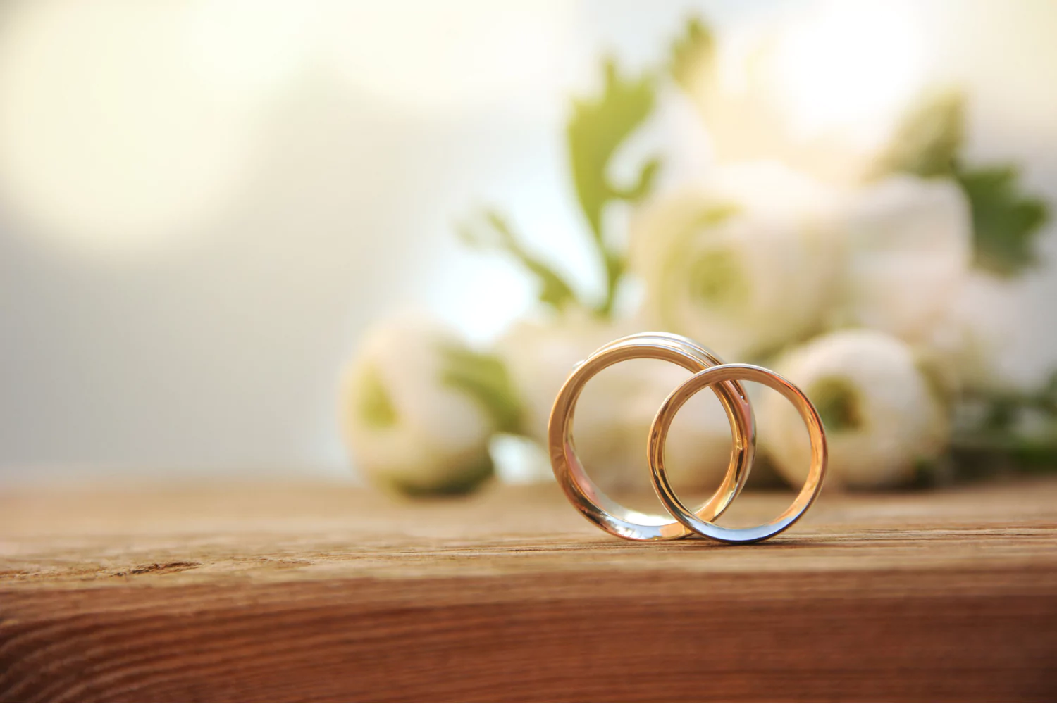 Two gold wedding rings on a wooden surface with white flowers in the background, symbolizing prioritizing marriage and lifelong commitment.