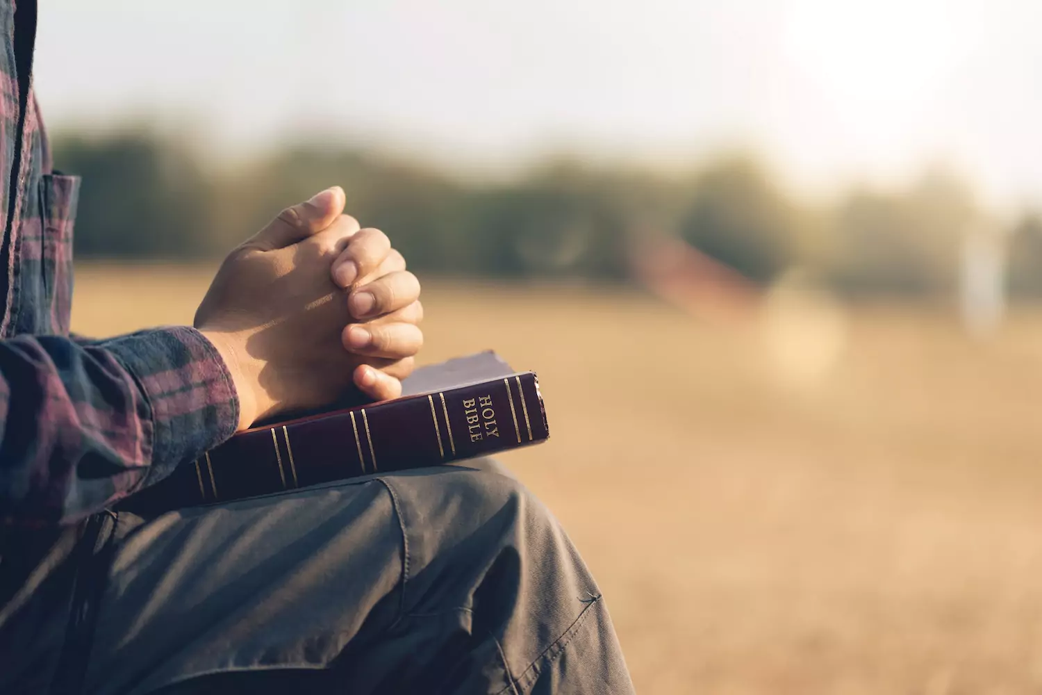 A man is sitting peacefully outside with a bible on his lap, and his hands folded in prayer, resting on the Bible.