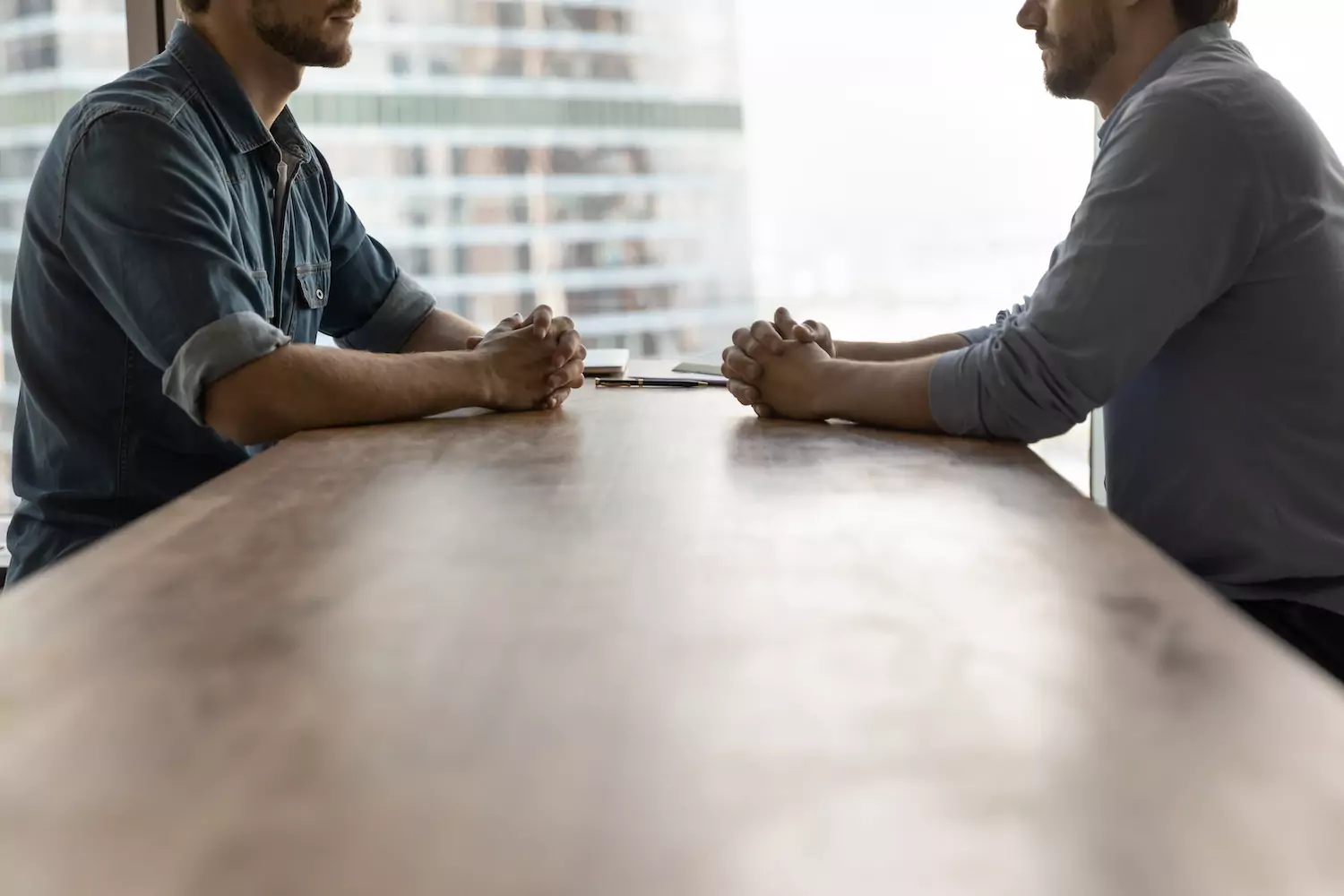 Two men sit at a table, face to face with each other, staring at each other in conflict.