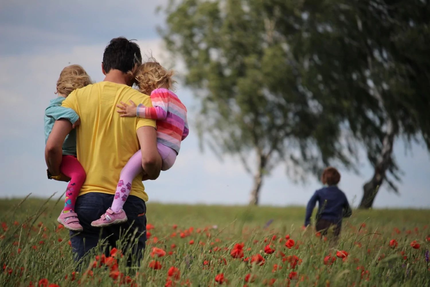 father holding his two children, one in each arm, as he follows another through the grass