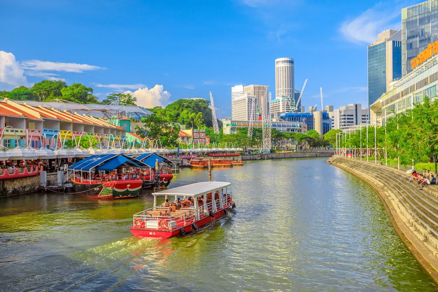 A photo of a city in Southeast Asia, with a boat floating down a river toward some large building in the background.