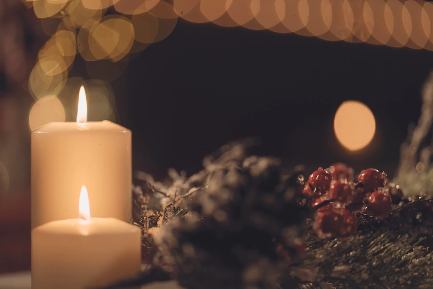 a close-up of two lit candles nestled among pine cones and holly