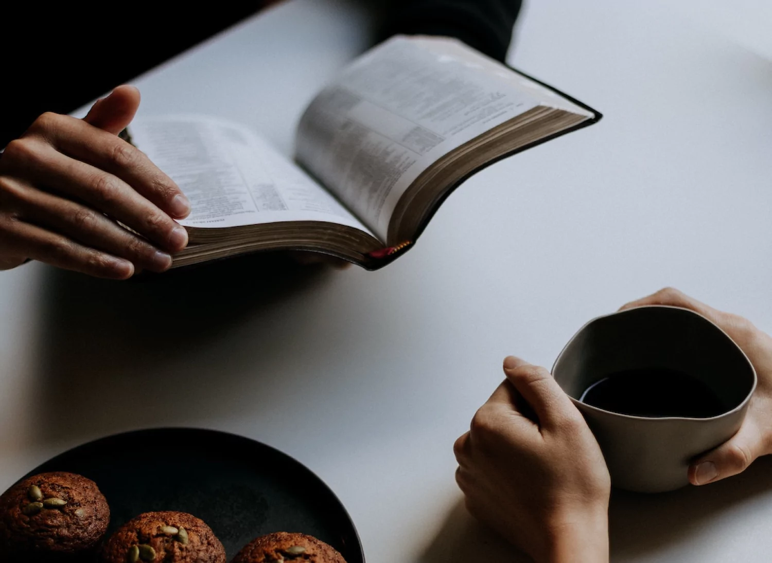 a premarital counselor with open bible in hand sits across from an engaged person holding a coffee mug