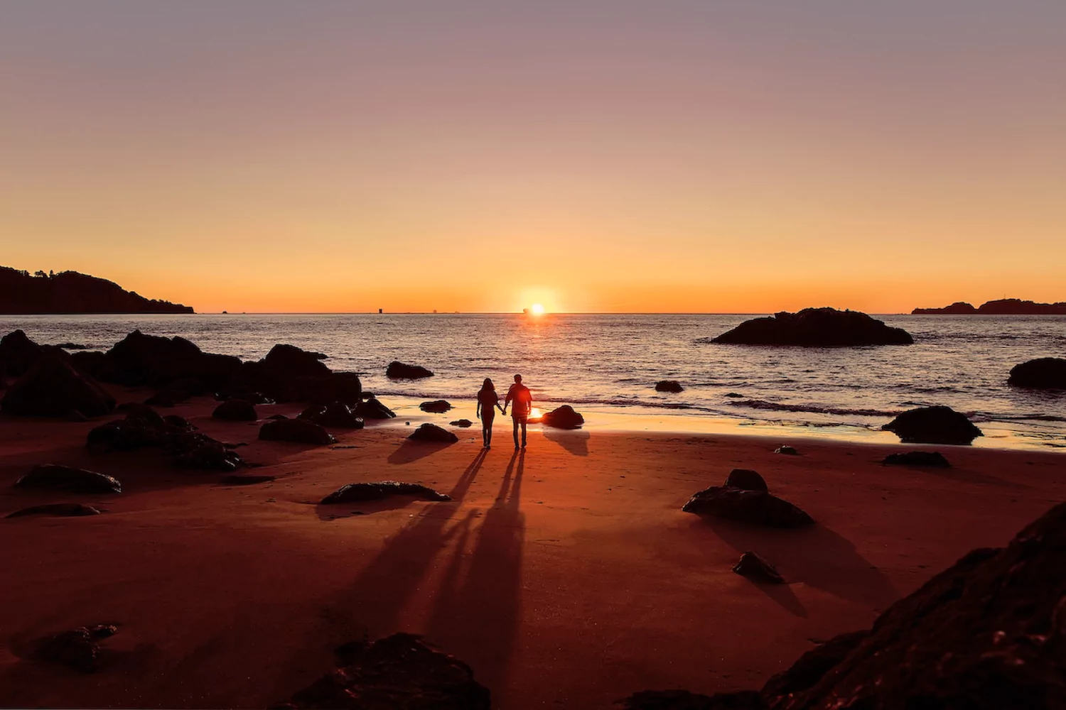 a husband and wife walk hand-in-hand at the beach at sunset
