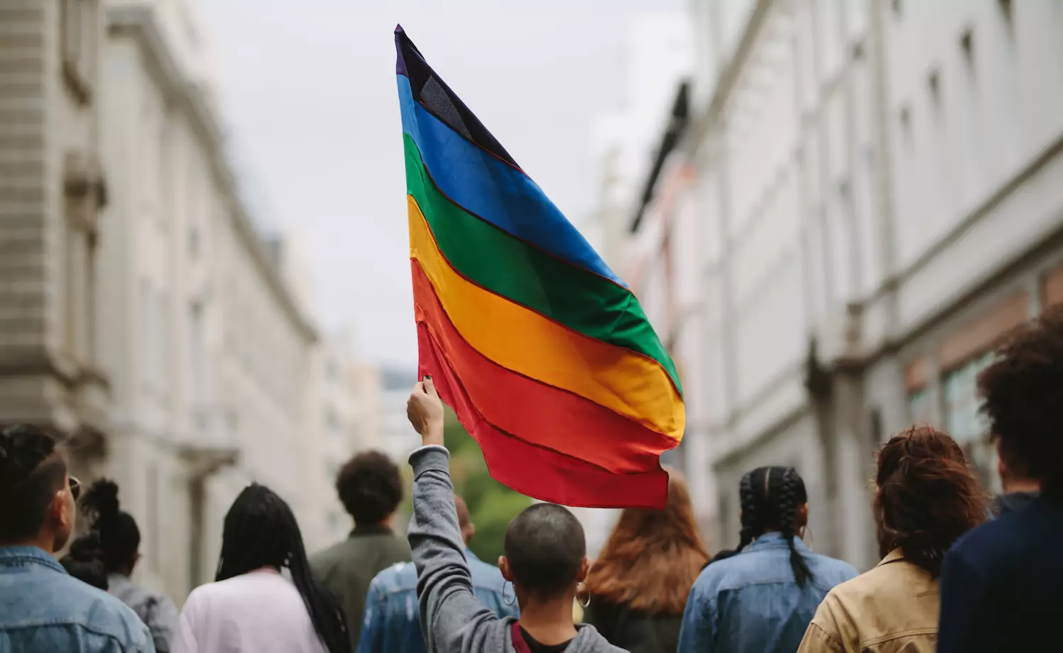 LGBT pride flag raised above a crowd on city streets, a broken symbol of the sexual revolution of our day.