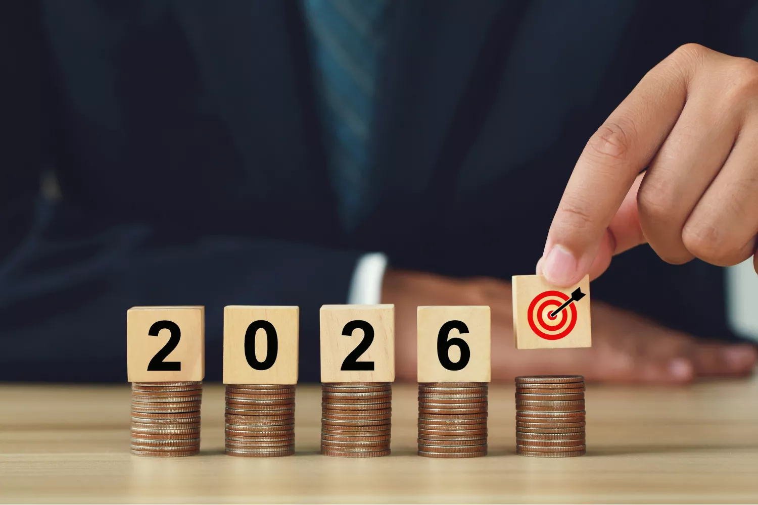Wooden blocks displaying “2026” sit on stacks of coins as a hand places a target icon block, symbolizing planning and setting new year goals.