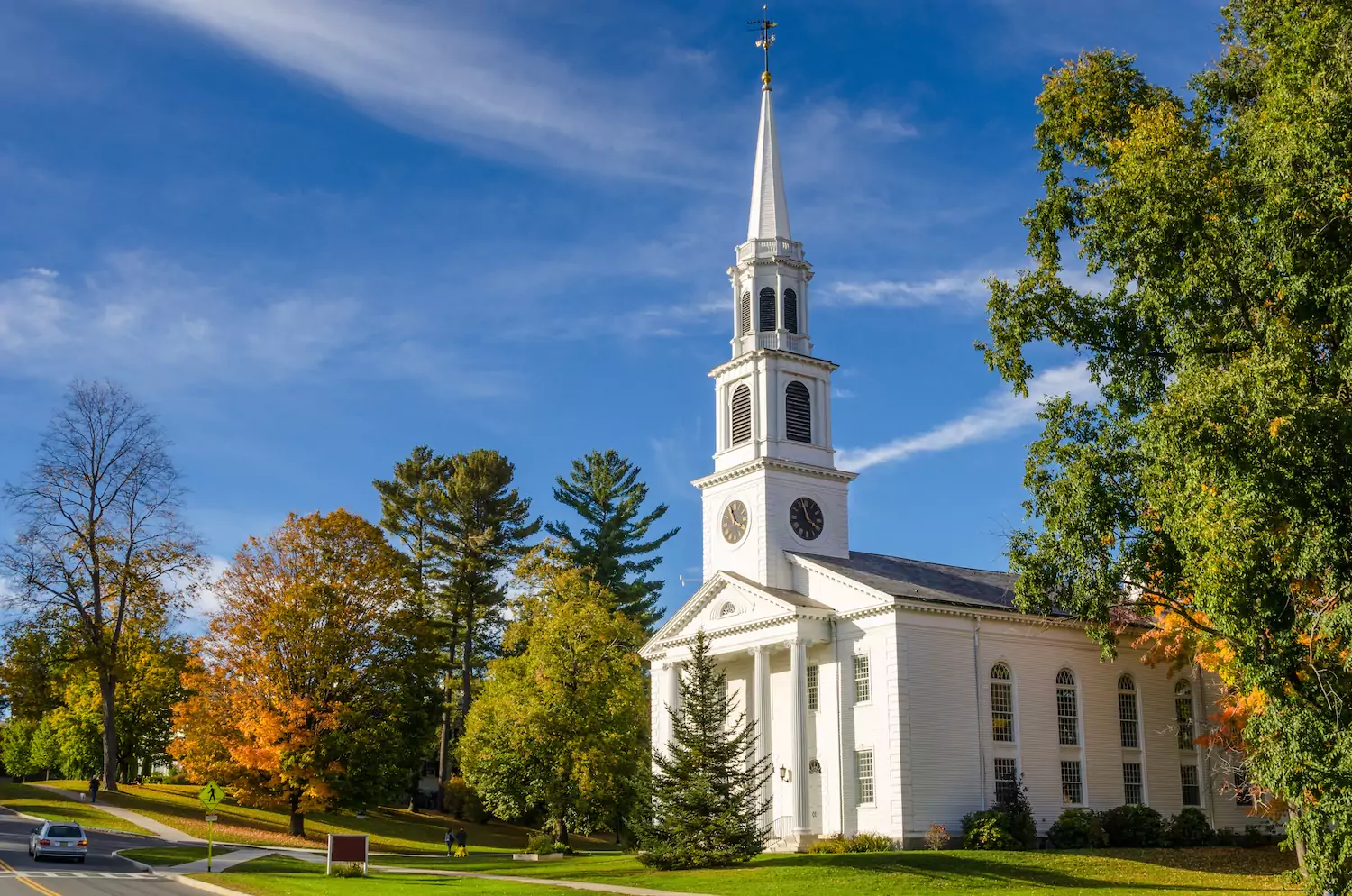 A white church with columns and a steeple stands among trees beneath a blue sky.