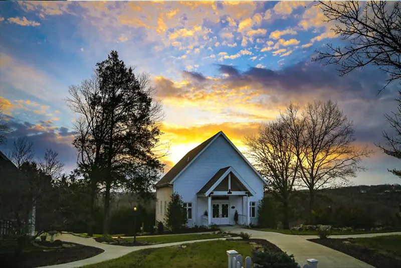 A picturesque white chapel at sunset, surrounded by trees and a well-maintained landscape with a winding path leading to the entrance.